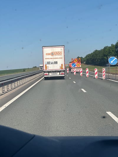 The image depicts a view from inside a vehicle, looking at the back of a KM Cargo truck, traveling on a highway with road work underway. The sky is blue, and the road is gray. The KM Cargo truck has the website address WWW.KM-CARGO.COM and the license plate number "02 AFQ 09" printed on the back. Road work is indicated by red and white striped barriers and road signs including a speed limit of 50. The location is not specified, so I cannot determine if it is in Poisevo, Russia.