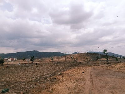 The image displays an expansive, semi-arid landscape located in Rafin Pa, Nigeria, under a heavily overcast sky. The foreground consists of dry, reddish-brown earth with sparse vegetation and a distinct dirt track originating from the lower right and extending into the midground. In the midground, several grey utility poles with light fixtures are erected, interspersed with scattered, small trees featuring green foliage. To the left midground, a cluster of multi-story buildings with light-colored facades and reddish-brown roofs is visible. Two individuals, clad in light-colored clothing, are standing near these structures. Approximately 50 meters to their right, a single individual, dressed in dark clothing, is observed walking away from the camera's perspective. The background features a continuous range of dark, undulating hills or low mountains stretching across the horizon. The sky is entirely covered by dense, grey and white clouds. A small, dark avian object is discernible in the upper right quadrant of the sky.