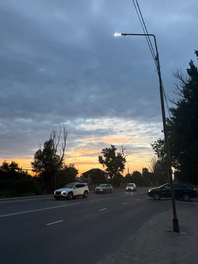 A street scene at dusk in Dachi, Kazakhstan, features a road with several cars. A white SUV is in the left lane, a silver sedan is in the middle lane, and a dark SUV is in the right lane. The sky above is filled with dramatic, dark clouds with streaks of orange and yellow from the setting sun. A street lamp with its light on illuminates the scene from the right side of the frame. Trees line the side of the road, and houses are visible in the distance. The overall impression is one of a calm, everyday moment on a road at the end of the day.
