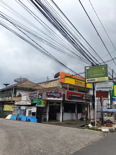 This image depicts a commercial street scene in what is described as Palayan, Philippines, under an overcast sky. The foreground features a gray asphalt road with a curb on the right.
Along the right side of the street, a cluster of multi-story buildings of varying architectural styles and conditions is visible. The leftmost building has a traditional brown tiled roof and a second-story veranda, where one individual in a blue shirt is observed, appearing to be working or painting on a wall. To its right, another building with a similar tiled roof displays multiple banners, including "Djarum Super" and "GrabFood." Further right, a more modern structure with yellow and orange upper levels features a "Yomart" sign and another "Djarum Super" advertisement. Two large, gray satellite dishes are mounted on the rooftops of the buildings.
Numerous black electrical or telecommunication wires crisscross extensively across the upper half of the frame against the cloudy sky. A prominent green sign on the right, supported by a utility pole, reads "CHOLOCO" and "EVERYONE CAN EAT CHOCOLATE," accompanied by text in Indonesian. Below this, another sign advertises "PERUMAHAN PORTIA ALAM GEULIS." A light-colored vehicle is partially visible parked on the far right, behind a street vendor's cart or stall. The overall lighting is dim, consistent with an overcast day.
