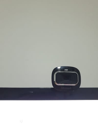 A black Microsoft webcam is positioned on a dark, reflective surface. The webcam has a silver trim around the lens and the "HD" logo is visible above the lens. The background is a plain, light gray wall. The overall scene is indoors, with no indication of the specific location in Proyonangg, Indonesia.