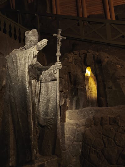 The image captures a dimly lit interior, likely a cavern or underground space, featuring religious sculptures. In the foreground, a large, gray stone statue of a robed figure resembling a bishop stands prominently, holding a cross in one hand and extending the other in a gesture of blessing or greeting. The statue is intricately carved, showing details in the robes and facial features.
In the background, another stone statue of a similar robed figure is illuminated with a warm, yellow light, creating a stark contrast against the dark stone walls. The surroundings appear to be rough and uneven, suggesting a natural or partially natural environment, possibly a cave or mine.
A wooden structure with decorative railings is visible in the upper portion of the frame, hinting at an elevated level or platform. The overall atmosphere is solemn and contemplative, possibly within a church or religious site, specifically located in Wieliczka, Poland.
