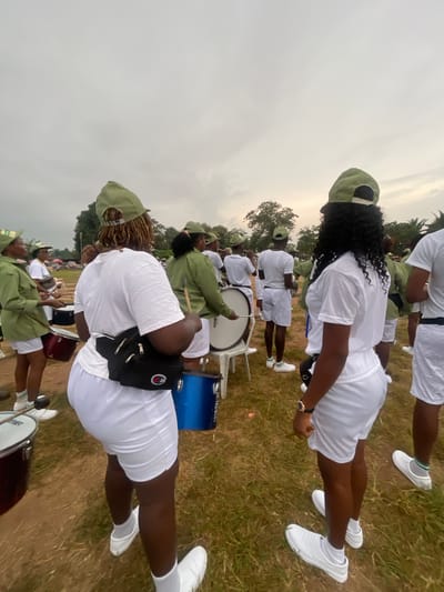 The image shows a group of young people, likely participants in a parade or performance, standing on a grassy field in Emure-Ekiti, Nigeria. The sky is overcast, suggesting it may be a cloudy day. The participants are dressed in matching uniforms, primarily white t-shirts and white shorts, with light green caps. Several individuals are holding or interacting with drums, indicating a drum corps or marching band activity. Some are holding drumsticks. The overall atmosphere appears to be one of preparation or performance. The terrain is uneven and covered with grass and some dirt. In the background, there are trees and other individuals, suggesting a public gathering or event.