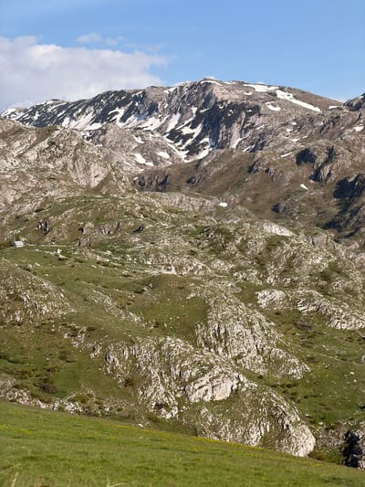 The image shows a mountain in Pišče, Montenegro. The left side of the mountain is rocky with patches of snow. The right side is covered in green grass and vegetation, mixed with rocky outcrops. A few small buildings are visible on the mountainside. The sky is light blue with some clouds.