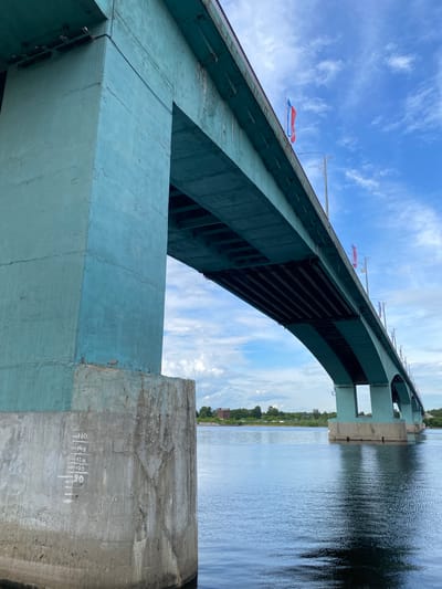 The image presents a low-angle view of a large arched bridge spanning a body of water, likely a river, under a bright, partly cloudy sky. The bridge's main structure is a robust arch, painted a faded light blue or teal, showing signs of age and weathering with some cracks and discoloration.

The perspective is from near the water level, looking upwards towards the underside of the bridge, revealing its structural beams and supports. Along the top edge of the bridge deck, numerous flags are visible fluttering in the wind, predominantly featuring red, white, and blue colors, consistent with the Russian national flag.

To the right, a wide expanse of water shows ripples and reflections of the sky and the bridge. In the distant background, a green shoreline with trees and a few buildings can be seen.

A large, unpainted concrete pier, supporting the bridge, is prominent in the lower right foreground. This pier exhibits white painted markings: "160", "140", "120", "100", and "90", each with a horizontal line, likely serving as water level indicators. The lower part of the pier has a darker, grittier texture, suggesting prolonged exposure to water.

The time of day is bright daylight, indicated by the clear blue sky and strong illumination. The overall scene suggests a functional bridge in a scenic, open environment, with clear skies. The presence of Russian flags aligns with the location context of Grigor'evskoe, Russia.