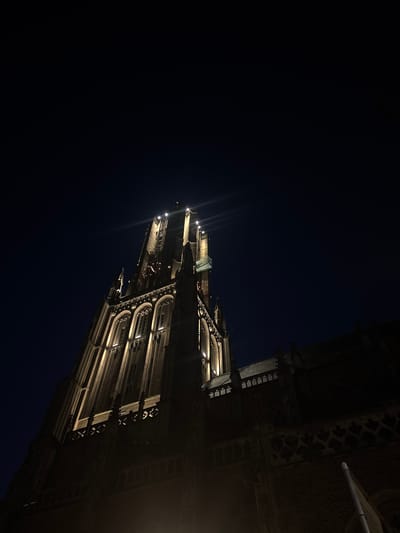 The image shows the tower of the Eusebius Church in Arnhem, Netherlands, at night. The church is illuminated by spotlights, highlighting its Gothic architecture. The sky is dark and clear. The perspective is from below, looking up at the tower. There are no people or animals visible in the image.