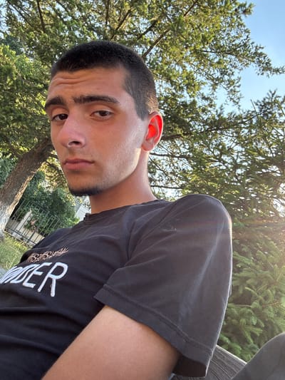 A young male with dark hair and a short haircut is pictured outdoors. He is wearing a black t-shirt with white lettering. The man is seated and looking towards the camera with a neutral expression. The background consists of green trees and a clear blue sky. The image appears to be taken during daylight, with sunlight casting a warm glow and lens flare in the upper right portion of the frame. The setting suggests an outdoor recreational area.