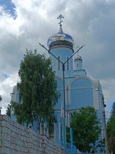 The image shows a bright blue church with a silver dome, topped with a cross. The church is partially obscured by a large green tree in the foreground. A brick wall is visible in the lower left corner of the image, and a street lamp is positioned in front of the church. The sky is partly cloudy with patches of blue sky visible. The overall scene appears to be a sunny day in a residential area. The church is located at 15 Sovetskaya Street, Yefremov, Tula Oblast, Russia.