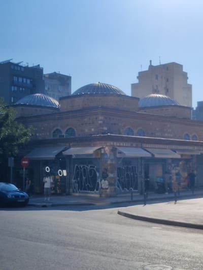 The image captures a street scene in Thessaloniki, Greece, on a clear, sunny day. The focal point is a historic building with multiple domes, constructed from brick and stone. The building features arched windows and a row of shops beneath awnings. The shops' windows and shutters are covered in graffiti.

In the foreground, a section of a road is visible, with a car parked on the left side. A person in a white t-shirt and shorts stands near the entrance of one of the shops. There are also several poles and a red circular sign with a white horizontal bar. In the background, modern buildings rise above the historic structure, indicating a blend of old and new architecture. The sky is a bright, cloudless blue, suggesting it is likely midday or early afternoon. The overall impression is of a lively urban environment with a rich historical backdrop.