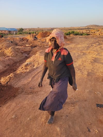 The scene is an outdoor, arid landscape in the city of Shen, Nigeria, characterized by reddish-brown earth, indicative of a disturbed area such as a quarry, brick-making site, or construction zone.
**Central Figure:** A dark-skinned individual, likely female, walks on an unpaved dirt path, moving towards the viewer. The individual is barefoot, and their skin, particularly on the arms and lower legs, is coated in reddish-brown dust. They wear a dark grey short-sleeved collared shirt with red shoulder and placket accents. A light-colored fabric, possibly faded pink, covers their head, draped to form a wide brim around the face. A dark skirt with faint vertical stripes extends below the knees.
**Environment and Background:** The ground is uneven, comprised of dry, compacted reddish-brown soil, with numerous excavated piles and depressions. In the mid-ground to the left, a single-story structure with reddish-brown walls and a light blue roof is visible, adjacent to a smaller, light-colored building. Distant, indistinct figures are scattered among earth mounds in the far background. Sparse green vegetation is visible beyond the immediate disturbed area. The sky is clear and light blue, suggesting fair weather. The overall lighting indicates either early morning or late afternoon.
