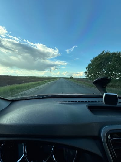 The image shows a view from inside a vehicle driving on a paved road. The road stretches into the distance towards a horizon, flanked by fields on either side. A tree is visible to the left of the road. The sky is blue with scattered clouds. A car dashboard is visible on the right side of the image. The location cannot be determined to be Merinovshchina, Russia based on the information provided in the image. There are no people visible.