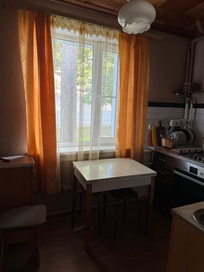 The image depicts an indoor scene, likely a kitchen or dining area, with natural light filtering through a window. The room appears modestly furnished and possibly dated.
In the foreground, a white-topped table with wooden legs is centrally placed. Two dark stools are tucked underneath the table. To the left, a portion of a wooden cabinet or dresser is visible with a drawer handle and a wooden stool with a textured seat.
The window, covered by orange curtains on the left and right and a sheer, lacy curtain in the center, overlooks an outdoor area with trees. A radiator is positioned below the window.
In the background, on the right side of the image, a kitchen counter is visible with a stovetop, sink, and various kitchen items such as dishes, cutting boards, and utensils. The walls appear to be painted a neutral color, and the ceiling is made of wood planks. A white, cloud-shaped light fixture hangs from the ceiling.
The overall impression is that of a simple, lived-in domestic space. The image does not contain any individuals or explicit actions.
Given the provided context, this scene is situated in the city of Naro-Fominsk, Russia.