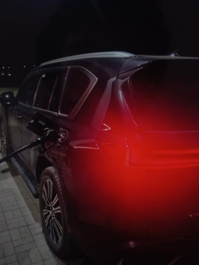 This media file shows the underside of a dark-colored car, likely at night given the lack of ambient light. The car is elevated, possibly by a lift or jack, revealing its undercarriage and a portion of its wheel. A bright red light emanates from the upper right portion of the image, casting a glow on the car's body. There is a noticeable vent or grille on the side of the car's body. The wheel visible has a multi-spoke rim with a silver and black pattern. The ground beneath the car appears to be paved with light-colored rectangular tiles. There is a small black object with some text on it, attached to a dark pole-like structure beneath the car. The scene suggests maintenance or inspection of the vehicle. While the location is specified as Yorkatay, Uzbekistan, there are no overt visual cues in the image to confirm this specific location. The time of day is clearly night. There is no indication of weather conditions or any people or emotions. The text on the small black object is not clearly legible.