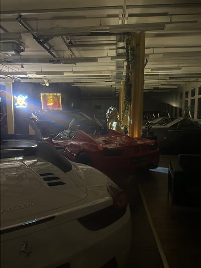 The image captures a scene in Wetzikon, Switzerland, featuring several luxury vehicles. A white Ferrari prominently occupies the foreground on the right side. Adjacent to it is a red Venturi, and further in the background is a dark-colored luxury car. The cars appear to be in a showroom or garage setting. The background shows illuminated signs, including one with "FRATTE LEONE" and another with a lion's head, suggesting the location is related to a business. The surrounding environment appears to be well-lit, with industrial-style elements such as pipes and overhead structures visible along the walls.