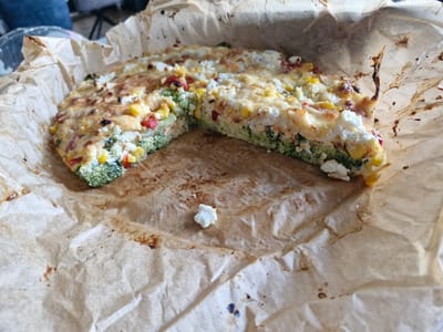 A close-up photograph captures a partially consumed, savory baked dish, resembling a vegetable quiche or frittata, resting on crumpled, grease-stained parchment paper. The dish, approximately semicircular with a significant wedge removed, displays a visible composition of green broccoli florets, yellow corn kernels, red bell pepper pieces, and white crumbled cheese, topped with a melted, lightly browned cheese layer. A solitary white cheese crumb is situated on the parchment paper in the foreground. The background is out of focus, revealing a portion of a person's leg in blue denim in the upper left quadrant, indicative of an indoor, domestic environment. The scene is located in Niedernhausen, Germany. No direct human action or interaction is depicted within the frame.