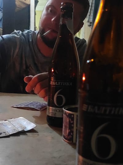 The image captures a close-up, slightly blurry scene at a table, likely in Makhinjauri, Georgia, given the provided context. In the foreground, two dark glass beer bottles are prominent, with the one on the left partially obscuring a man. The man, seen in profile with his back to the camera, is smoking a cigarette. He has a beard and is wearing a grey t-shirt. His right hand is visible, reaching towards the table. Lying on the table are several playing cards, fanned out and partially covered by what appear to be banknotes. The bottle on the left has a label with Cyrillic text and the number "6" prominently displayed, suggesting it's a "Baltika 6" beer. The background is dark and indistinct, with hints of light sources and what might be a doorway or window. The overall ambiance suggests a casual, possibly late-night setting.