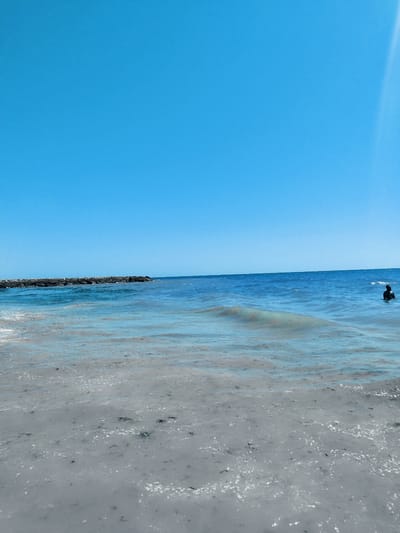 The image shows a clear blue sky over a calm ocean. In the distance, a man is partially submerged in the water, facing away from the camera. To the left, a rocky breakwater extends into the sea. The water is a vibrant blue, and the shallow area near the shore is a light grey with small dark debris scattered across the surface. The sun is shining brightly, casting a gentle ripple pattern on the water. The scene evokes a sense of peace and tranquility, suggesting a warm, sunny day at the beach in Rota, Spain. There is no visible text in the image.