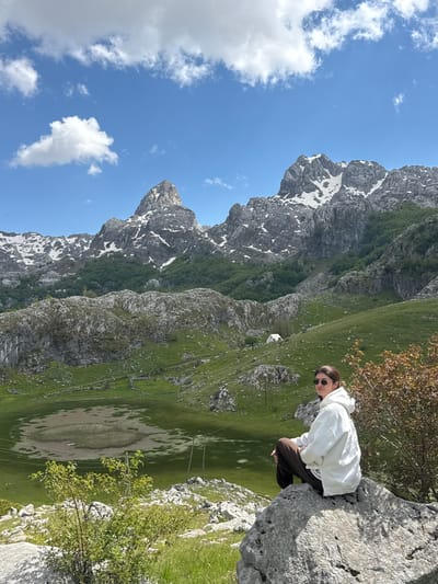 A slightly rotated, eye-level outdoor shot in Brskut, Montenegro, shows a person sitting on a rocky outcrop overlooking a valley with snow-capped mountains in the background, under a blue sky with scattered clouds.

The person is wearing sunglasses, a white hoodie, and dark pants, and is crouched on a light grey rock formation on the right side of the frame. They are looking toward the valley, which is lush with green grass and contains an oval shaped, dried up or shallow lake or depression. A few small, indistinct buildings are visible in the valley. Power lines extend vertically across the valley.

On the left side of the frame is a mountain range with grey, rocky peaks speckled with snow. The sky above is a bright blue with scattered white clouds. The perspective is that of a wide shot, showing a panoramic view of the mountainous terrain.