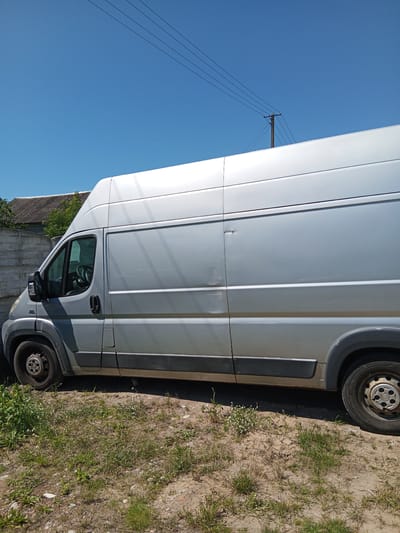 A silver panel van is parked on a dirt and grass area next to a concrete wall. The van is facing left, and its side door is visible. The sky above is clear and blue, with power lines and a utility pole in the distance. The background shows a tiled roof and trees. The scene appears to be in the city of Leoncin, Poland.