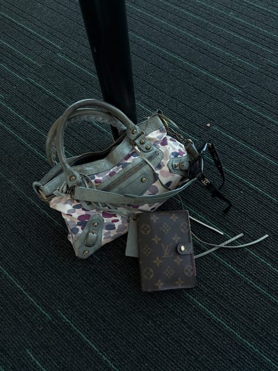 The image depicts a still life arrangement of personal belongings against a dark carpet with subtle green linear patterns. A handbag, predominantly gray with a printed fabric accent featuring purple and white blotches, lies open, revealing its interior. Atop the handbag rests a pair of black Chanel sunglasses. To the right of the handbag, a brown Louis Vuitton wallet is positioned. The background is dominated by the dark carpet, which appears to be tightly woven. A portion of a black, cylindrical object, possibly a bar or pole, is visible towards the left side of the frame. The scene appears to be indoors. There is no information to make a determination about the location in Uttlesford, UK.