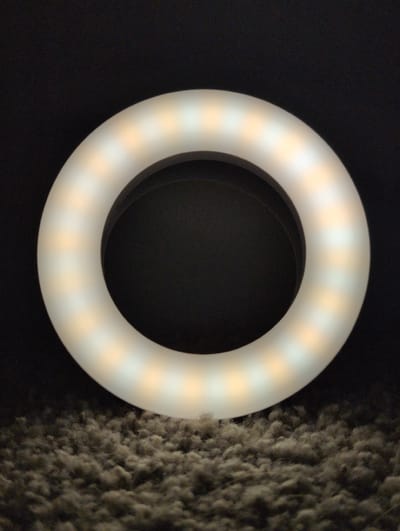The image shows a close-up, high-angle shot of a white ring light with yellow and white LED lights, placed on a dark, fuzzy surface that appears to be carpet or fabric. The ring light is the primary subject, positioned in the center of the frame. The background is dark and out of focus. The lighting from the ring light casts a glow, creating a circular illumination effect. The overall scene is static and focuses on the object and its immediate surroundings. There are no people or discernible landmarks of Louisville, United States of America visible in the image.