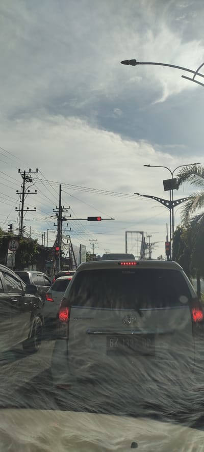The image depicts a traffic jam on a cloudy day in Kuta Lhokseumawe, Indonesia. The view is from inside a vehicle, looking forward through the windshield and the rear of the car in front. Several vehicles are visible, including the white car directly ahead with a license plate that reads "BK 133 OP". Traffic lights are red, indicating a stop. Utility poles with numerous wires are prominent features of the urban landscape, along with street lights and palm trees. The overall scene suggests a typical urban environment experiencing traffic congestion.