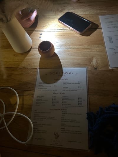 The image shows a tabletop in a bar or restaurant, with a menu titled "Doki Doki Cantina" visible. There is a small ceramic mug, a cell phone, and a white vase with dried flowers in the top left corner. The surface is made of wood and is lit by an overhead light. In the bottom right corner, a blue cloth is partially visible. There is a cactus logo printed on the bottom of the menu. The location of this image is unknown as coordinates do not provide information about the location of a specific image.