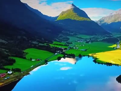 This is an aerial, high-angle drone shot of a picturesque rural landscape. The scene is dominated by a large body of calm, blue water in the foreground, reflecting the sky and clouds. The water's edge is lined with a verdant embankment, with some dark green trees. Beyond the water, a lush green valley unfolds, dotted with small buildings, including red and white structures that appear to be farmhouses or barns. A road winds through the valley, with a few white vehicles visible. The valley is framed by imposing, dark blue mountains that recede into the distance. One prominent, conical mountain peak in the mid-ground is illuminated by sunlight, casting a bright green and yellow glow on its grassy slopes. The sky is a vibrant blue with scattered white clouds. The location is identified as Olișcani, Moldova.
