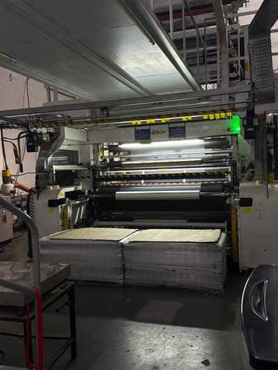 This image captures an interior view of an industrial manufacturing facility in Gamovo, Russia. The scene is dominated by a large, complex machine featuring multiple rollers, metal frames, and various components indicative of a production line, possibly for printing, converting, or processing sheet materials.
In the center, large, metallic rollers are prominent, with bright artificial lighting illuminating the area. A distinct green indicator light is visible on the upper part of the machinery, suggesting operational status or a system alert. To the right, stacks of what appear to be processed sheets or raw materials, possibly paperboard or similar goods, are neatly arranged and wrapped in plastic. The floor is concrete with visible cracks and stains, typical of an industrial environment. Overhead, an intricate network of metal structures, conduits, and wires forms part of the facility's infrastructure.
Visible text on some panels or boxes in the upper right part of the machine is in Russian. One reads "ПРОВЕРЬ РАБОТУ АНТИСТАТИЧЕСКИХ ПЛАНОК" (Check the operation of anti-static bars) and another "ПРОДАТЬ АНТИСТАТИЧЕСКИЕ ПЛАНКИ" (Sell anti-static bars). A logo that appears to be "М-М" is also present. Yellow and black striped warning tape is wrapped around some structural elements. The facility is under artificial lighting, with no natural light sources visible, suggesting it could be any time of day or night. No people are present in the frame, but the machinery implies ongoing or readiness for industrial activity.