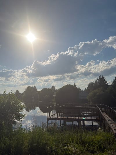 The image captures a serene natural scene with a body of water reflecting the bright sun and clouds. A wooden pier extends into the water, with a ramp leading to it. Lush green grass and trees surround the pier and the water's edge. In the background, houses are visible amidst the trees, suggesting a rural or suburban setting. The sun is high in the sky, casting a bright glare. The location appears to be Малое Седельниково, Russia, a village known for its natural beauty and peaceful atmosphere.