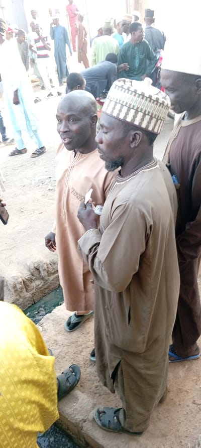 This image depicts an outdoor gathering of approximately twenty or more adult males in Kabuga, Nigeria. In the immediate foreground, three individuals are prominent. The leftmost subject is a bald male wearing a light beige long-sleeved garment with dark embroidery and dark open-toed sandals, exhibiting an open-mouthed expression and directing his gaze slightly right. Adjacent to him, a male with a short dark beard and mustache wears a light brown long-sleeved garment and a white and brown patterned cap, holding several white paper currency notes in his right hand at chest level, with his gaze directed downwards. To his right, a third male is partially visible, wearing a dark brown long-sleeved garment.

The background shows additional males in various traditional garments, including light blue, green, and white, suggesting a crowded public space. The ground surface is unpaved earth. A narrow, open concrete drainage channel containing murky liquid runs horizontally across the lower mid-ground. Footwear observed on visible individuals includes various sandals and flip-flops. The scene is illuminated by natural daylight.