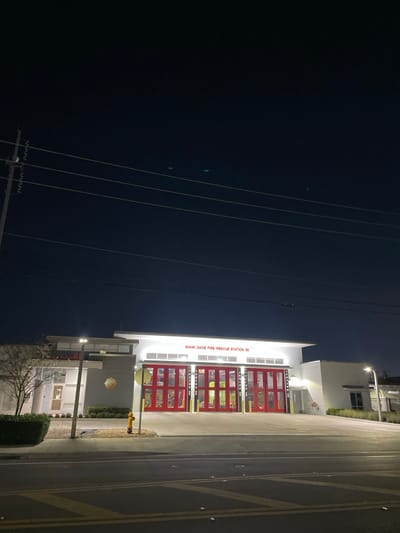 Miami-Dade Fire Rescue Station 66 at night shows a modern, two-story building. Three garage doors, illuminated in red, stand out. Streetlights cast shadows on the adjacent road, marked with yellow lane lines. Power lines traverse the dark sky. A yellow fire hydrant is visible near the station.