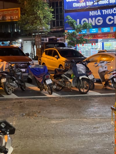 The image captures a street scene in Bắc Hà, Vietnam, at night. The foreground shows several parked motorcycles, some with blue covers. A dark-colored pickup truck is parked behind them, and to its right, a yellow vehicle, possibly a car or van, is also visible. In the background, a building with bright blue signage advertising "NHÀ THUỐC LONG CHI" (Long Chi Pharmacy) is prominently displayed. Other signs and store windows are illuminated, indicating a commercial area. The street appears wet, suggesting recent rain. The overall atmosphere is that of a typical urban street at night, with vehicles and commercial establishments lining the road.
