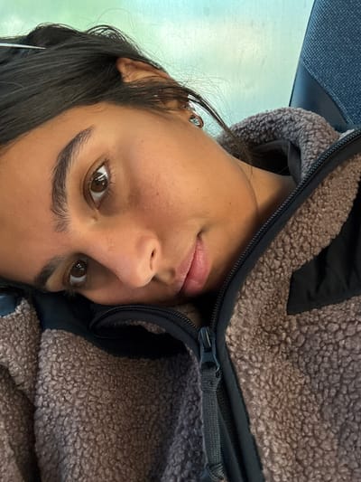 A close-up image depicts a young adult female from the chest up, leaning to her left, with her head resting on what appears to be a dark upholstered seat or headrest. She has dark hair, brown eyes, and a light to medium skin tone, with a neutral expression as she looks directly towards the camera. She is wearing a textured brown or tan fleece jacket with a visible black collar and a partially unzipped black zipper. Small, reflective earrings are visible on her left ear. The background is blurred, displaying a light blue-green hue, possibly indicating a window or interior lighting. The specific location within Linden, United States, cannot be determined from the visual information provided in the image.