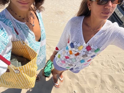 Two individuals stand on a sandy surface in Caminha, Portugal. The person on the left wears a white and turquoise patterned tunic over a dark top, multiple necklaces, and dark flip-flops, carrying a yellow and white striped tote bag with red handles. The person on the right, likely taking the photo from a high angle, wears a white crochet cardigan with multicolored floral appliqués, light-colored shorts, sunglasses, a delicate gold necklace, and white flip-flops, holding a small woven bag containing a green rolled item. In the upper right background, part of a vehicle with a visible license plate "2685 VF" is present. The scene is illuminated by strong sunlight, casting shadows on the sand.