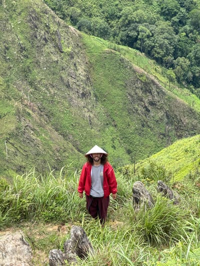 A person stands on a grassy hillside wearing a red jacket, a grey t-shirt, maroon pants, and a conical hat. The background features a lush green mountainous landscape with dense forest covering the upper slopes. The terrain is steep and covered in vegetation. The person is smiling at the camera, and their body is slightly angled. The overall scene depicts an outdoor adventure in a natural, hilly environment. This appears to be the highlands of Bắc Yên, Vietnam, characterized by its verdant, rolling hills and mountainous terrain.