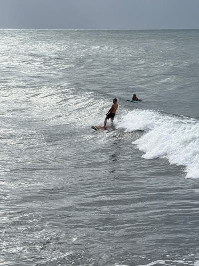 An outdoor wide shot captures two individuals in the ocean off the coast of Māhina, France, under an overcast sky. In the midground, a male with a beard, shirtless and wearing dark shorts, actively surfs a breaking wave from right to left on a light-colored surfboard with dark stripes. White foam forms along the crest of the wave he is riding. Further out in the background, a second individual, wearing a dark top, lies prone on a light blue surfboard, appearing to wait. The ocean surface exhibits ripples and reflects a dull silver-grey light. The sky above is a uniform light grey, blending with the horizon line.