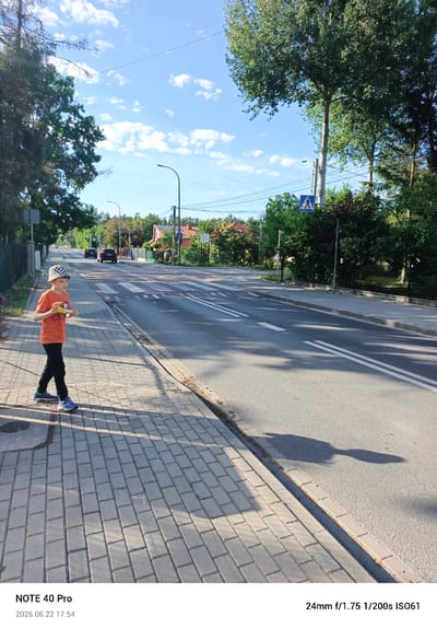 A young boy wearing a checkered hat, an orange t-shirt, and black pants stands on a paved sidewalk next to a road in Kamionka, Poland. He is holding a yellow object in his hands. The boy is looking towards the camera. The scene appears to be outdoors on a sunny day, indicated by the shadows on the sidewalk and the clear blue sky with scattered white clouds.

The road in front of him has a pedestrian crossing. Cars are visible in the distance on the road, and houses with red roofs are seen among the trees lining the street. A blue traffic sign depicting a pedestrian crossing is also visible. The overall setting is a suburban or rural road. The time of day is 17:54, indicated by the text at the bottom of the image. The date is June 22, 2026. The camera used was a NOTE 40 Pro with settings of 24mm f/1.75 1/200s ISO61.
