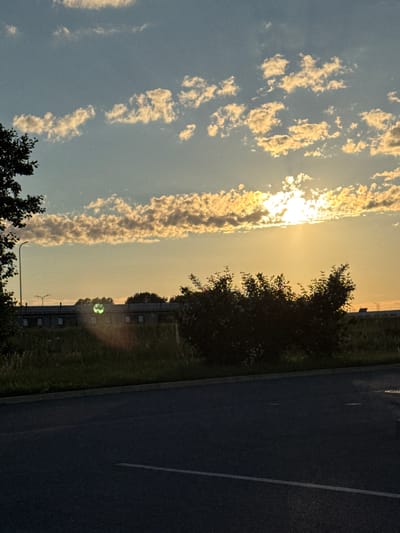 The image captures a late afternoon or early evening scene in Laagri alevik, Estonia. The sky is a soft blue, transitioning to yellow at the horizon, and is dotted with scattered, fluffy clouds that are illuminated by the setting sun. The sun itself is partially obscured by clouds, creating a bright glow with visible rays. In the middle ground, a long, low-rise building with several windows is visible, set against a backdrop of trees and vegetation. A lamppost stands to the left of the building. In the foreground, a dark asphalt road with a white line marking runs diagonally across the frame. The overall impression is one of tranquility and the end of a day.
