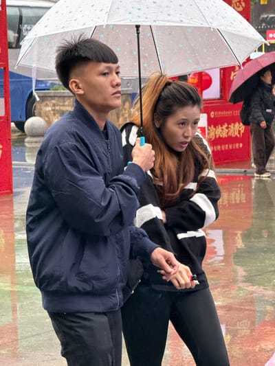 This image depicts an outdoor scene during rainfall in Pengshui Miao and Tujia Autonomous County, China. Two individuals, a male and a female, occupy the foreground. The male, positioned on the left, has short, dark hair with a faded side and a bowl-cut top. He wears a dark blue bomber jacket over a dark shirt and black pants. He holds a clear umbrella with small blue star patterns in his right hand, the handle of which is light blue. His gaze is directed forward. His left hand is clasped with the female's right hand.
The female, to the male's right, has long, reddish-brown hair tied in a high ponytail. She wears a black sweater with prominent white horizontal stripes and black leggings. Her mouth is slightly open, and her eyes are directed towards her left (viewer's right). In her right hand, which is held by the male's left, she holds a small red object, appearing to be a lighter.
The ground is wet and reflective, indicating recent or ongoing rain. In the mid-ground to the right, a third individual is partially visible, holding a dark red umbrella. Further in the background, red structures and signage are present, including text that appears to be "渝快茶消费" on a red surface. A portion of a blue vehicle or structure is visible on the far left of the frame.