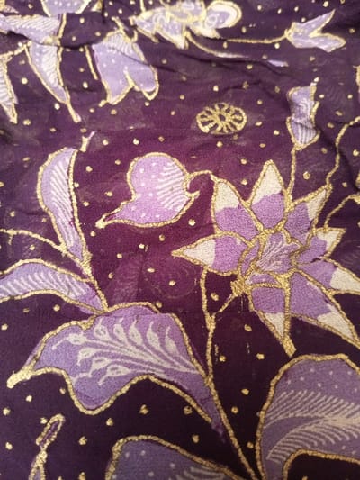 A close-up shot reveals a patterned fabric, predominantly dark purple with floral designs. The flowers and leaves are rendered in shades of light purple and lavender, outlined by fine gold lines. Small gold dots are scattered across the fabric's background. The texture appears soft and slightly translucent, suggesting a light material.