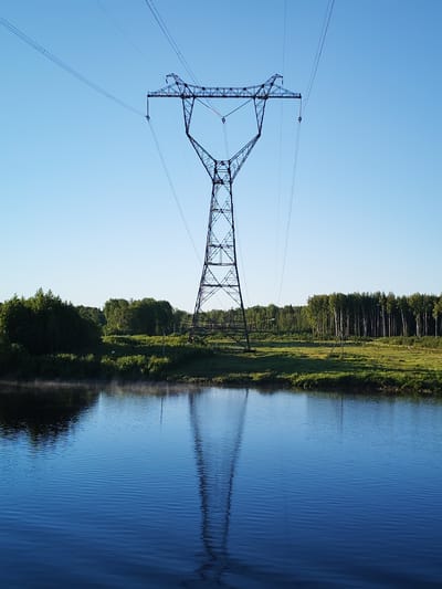 The scene is situated in the city of Gari, Russia. A large, dark-colored lattice steel electrical transmission tower stands centrally, rising from a vegetated midground and extending towards the top of the frame. Multiple high-voltage power lines, arranged in parallel bundles, are suspended from the tower's cross-arms and extend horizontally against a clear, light blue sky. The lower half of the image features a calm body of water that reflects the tower and sky, with minor surface ripples subtly distorting the reflection. A faint layer of mist is present just above the water's surface along the far bank, particularly on the left side. The land bordering the water consists of green grass and low-lying green vegetation. Beyond this foreground, a dense treeline forms the background, characterized by dark green, leafy deciduous trees on the left and a distinct stand of slender, light-barked trees on the right. A smaller wooden post is visible in the grassy area to the right of the main tower's base. The environment appears naturalistic. No individuals or animals are present in the image.