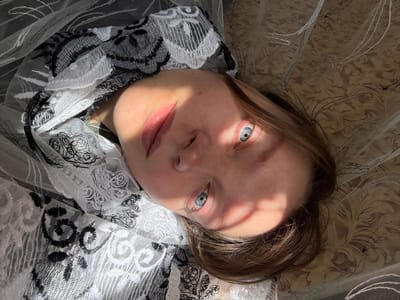 An eye-level shot captures a person, likely female, lying down and looking directly at the camera. The subject has fair skin, striking light blue eyes, and light brown hair that frames their face. Their expression is neutral and direct, with a subtle intensity in their gaze.

Strong, directional sunlight illuminates the scene, casting intricate geometric patterns of light and shadow across their face, particularly over the forehead and eyes. The person is partially draped or surrounded by translucent fabric, which appears to be a white and black lace or embroidered mesh material featuring distinct floral and scroll patterns. This fabric creates a soft, diffused boundary around the subject, with brightly lit sections highlighting its delicate textures. Another lighter, patterned fabric in warm, muted tones (beige/cream) forms the background, suggesting bedding or upholstery.

The overall setting points to an indoor environment, likely a bed or sofa. The intense sunlight implies it is daytime, possibly morning or late afternoon, with clear weather outside. The image focuses on the person's upper body and face, emphasizing their direct gaze and the interplay of light and shadow. The scene conveys a sense of stillness and contemplation. This media file was captured in Aksay, Kazakhstan. There is no visible text in the image.