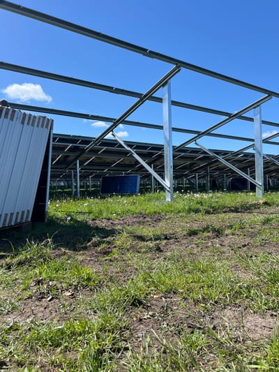 The image shows a ground-mounted solar panel installation under construction, likely in a rural or semi-rural setting near Grange Ville, Ireland. The foreground features grassy terrain mixed with exposed soil. In the background, rows of metal framework are visible, designed to support the solar panels. Some solar panels are already installed. A stack of white or light-colored panels or construction materials is visible in the bottom-left corner. The sky is clear blue with scattered white clouds.
