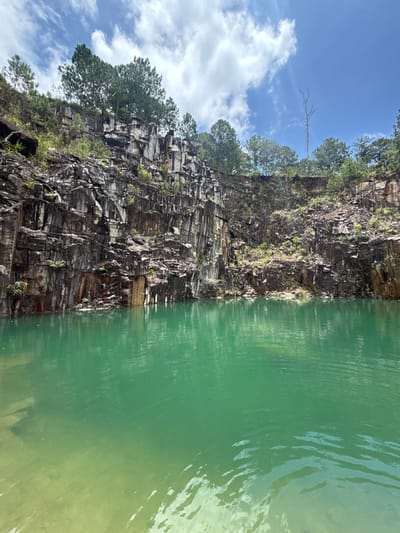 The image shows a quarry with sheer, rocky walls surrounding a body of clear, turquoise water. Pine trees grow on the rim of the quarry and in some areas along the rock face. The sky above is blue with scattered white clouds. The quarry is located in the city of Đăk Hơ, Vietnam. There are no people or animals visible in the image. The environment is natural and serene.