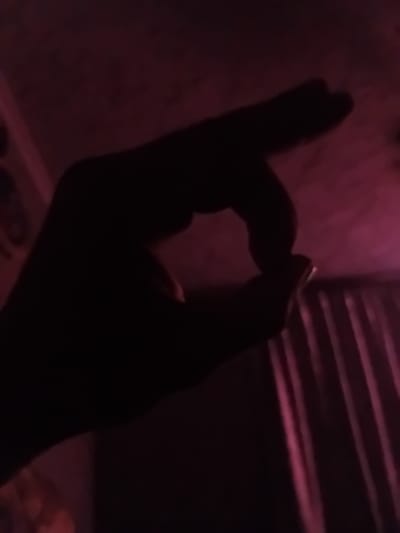 A silhouette of a hand forms an "O" shape against a dark, dimly lit background with pinkish hues. The hand is positioned in the foreground, with the thumb and index finger nearly touching, creating a circular opening. The background appears to be an interior space, possibly a room, with curtains visible on the right side. The lighting suggests an artificial light source, casting a subtle glow on the surroundings. The scene is devoid of any specific identifiable landmarks or context related to the city of Verkhniachka, Ukraine, and offers no discernible information about any individuals or actions taking place beyond the formation of the hand gesture.