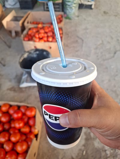 A hand holds a Pepsi cup with a blue straw in front of a background of tomatoes in cardboard boxes at an outdoor market in Ko`kdala, Uzbekistan. The cup is black with a blue dotted pattern and the Pepsi logo. In the background, several cardboard boxes are filled with ripe red tomatoes. To the left of the cup, a black, round object, possibly a watermelon, sits on a scale. The ground appears to be concrete or dirt.