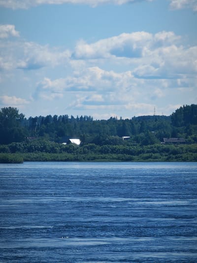 This is a vertical, eye-level, medium shot photograph of a wide, blue river under a partly cloudy sky. The foreground is dominated by the rippling water, which reflects the sky with horizontal lines of light and shadow. In the middle ground, a lush green bank is lined with trees and bushes. Scattered amongst the foliage are several buildings, including houses with pitched roofs and a white structure that gleams in the sun. The background features a dense forest that covers rolling hills, with a faint silhouette of a communication tower visible on the highest peak. The overall impression is one of a serene, rural landscape. The scene is consistent with the village of Абалаково, Russia, situated along the Yenisei River.