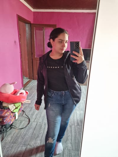 A woman is taking a selfie in a mirror. She is wearing jeans, a black t-shirt with "Los Angeles" printed on it, and a black jacket. Her hair is pulled back. She is holding a smartphone in her right hand, with the camera facing the mirror. The reflection shows part of a pink wall with a brown-framed window. The floor is tiled with gray wood-patterned tiles. There's a stuffed animal and a suitcase with wires/cords on the floor next to the mirror. The location information indicates this is in Byala Slatina, Bulgaria.