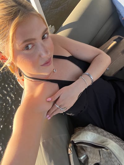 The image is a selfie-style shot of a woman with fair skin and light blonde hair. She is wearing a black top, several bracelets, and rings. She also has a choker and a necklace. To the side is a Gucci handbag. The photo seems to be taken on a boat, evidenced by the water in the background. The location cannot be verified as Livingstone, Zambia, from the image alone. No actions or interactions are visible. The environment includes the water and the boat's seating.